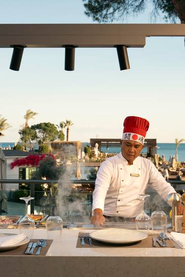 A chef in a white uniform and red hat cooks at an outdoor kitchen station at UMI Teppanyaki Restaurant, with steam rising from the grill and a scenic view of the ocean, palm trees, and resort guests in the background